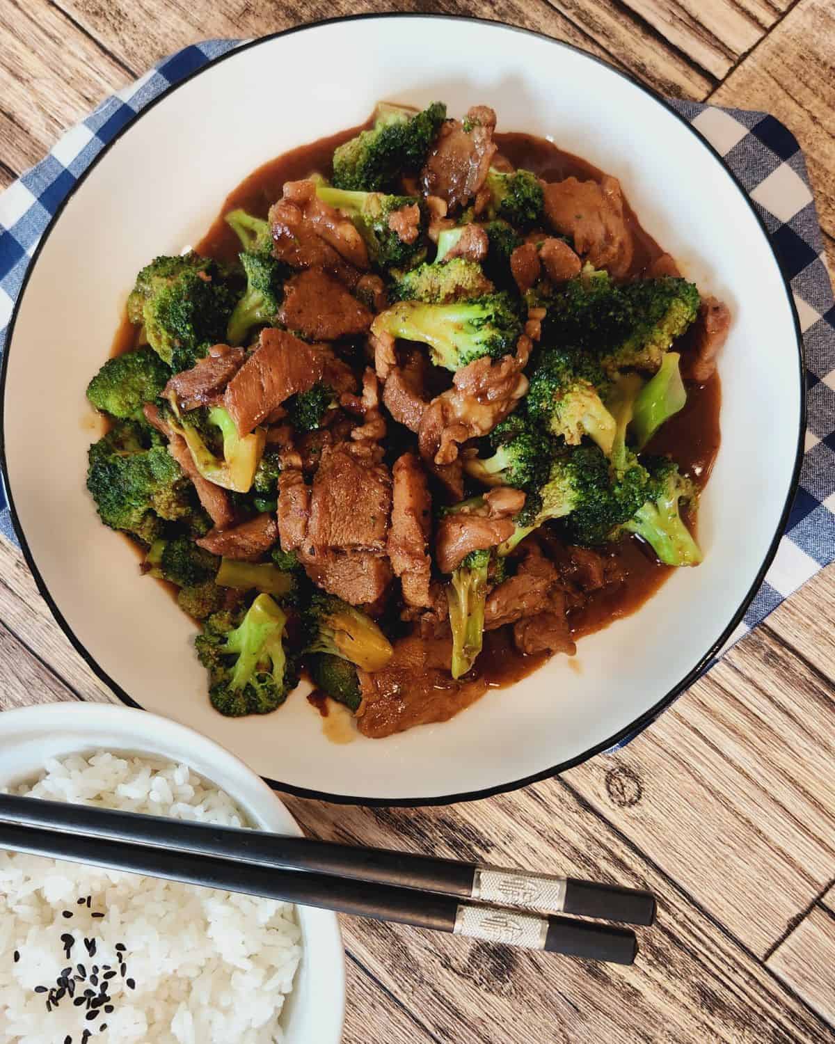 A plate of pork and broccoli stir fry with a bowl of rice and chopsticks.