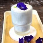 A glass of Butterfly Pea Flower Latte with some fresh flowers on a wooden tray.