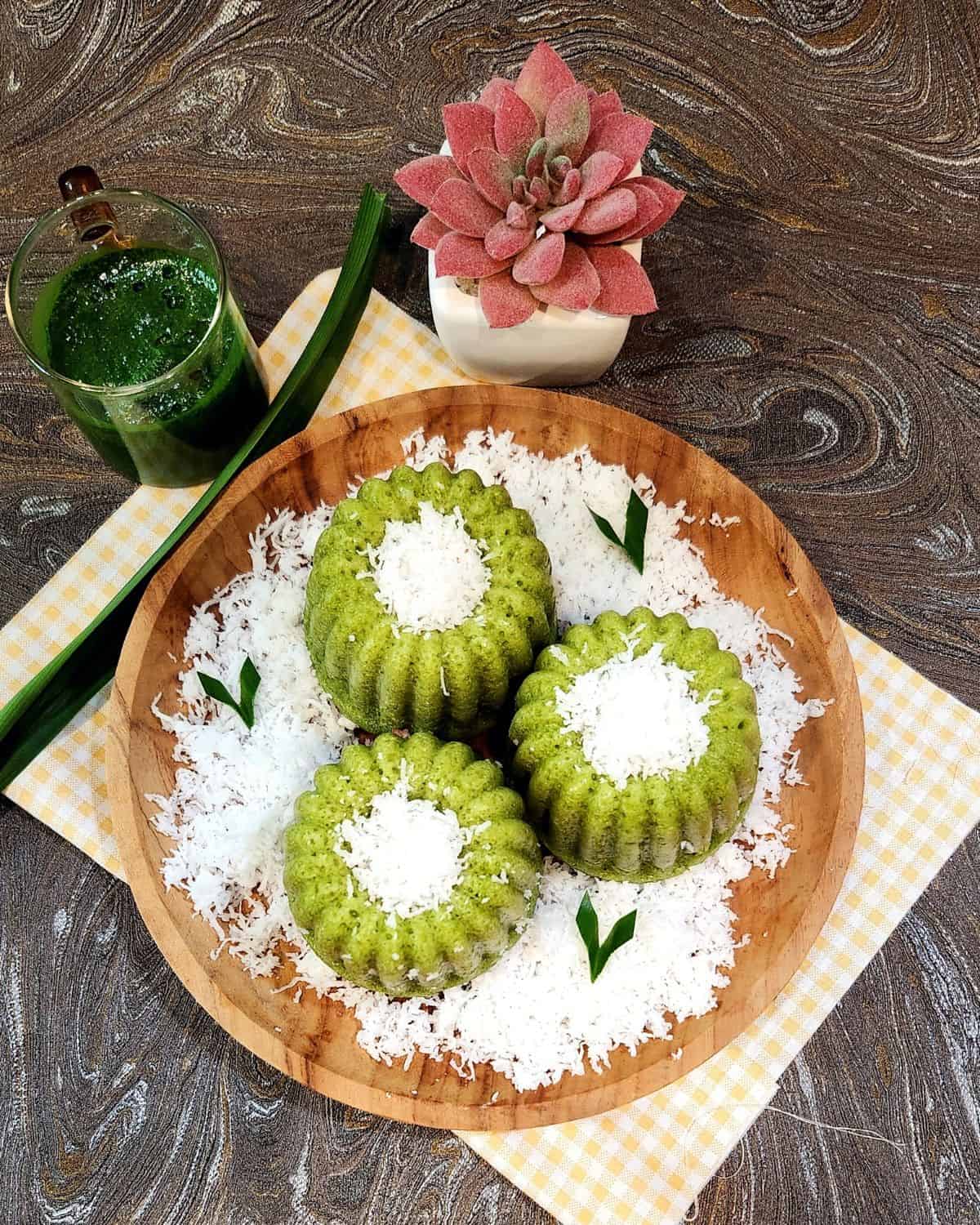 Three kue apem pandan on a wooden plate with grated coconut and pandan extract next to it.