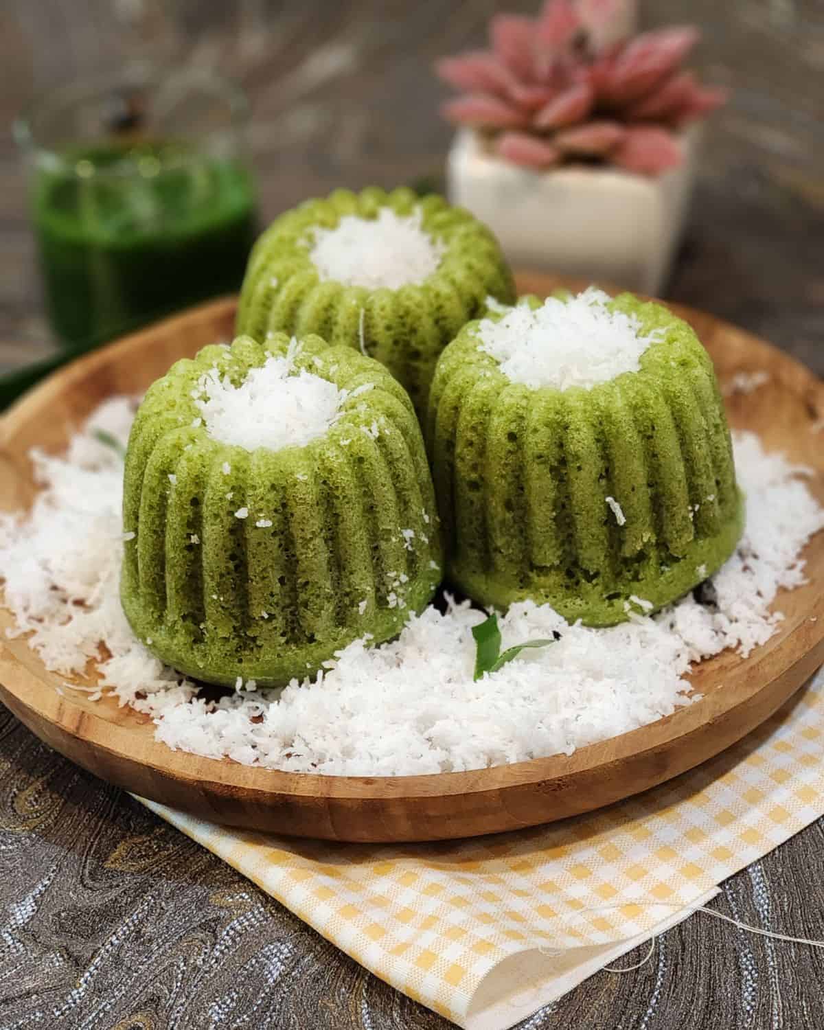 Three kue apem pandan on a wooden plate with grated coconut and pandan extract next to it.