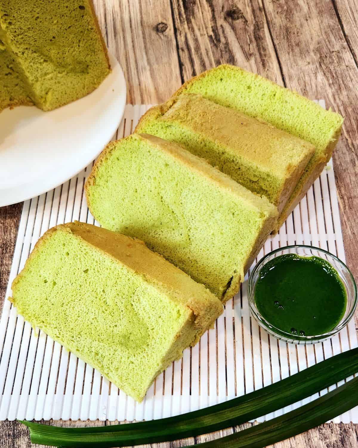 A few slices of pandan chiffon cake on white plastic tray with a cup of pandan extract next to it.