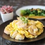 Chinese shrimp omelette on a black plate garnished with curly scallion on top, at the background there is another simple dish.