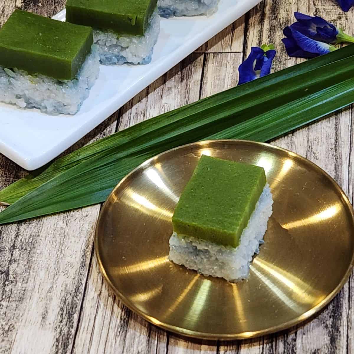 A golden plate of kuih seri muka with butterfly pea flowers and pandan leaves at the back.