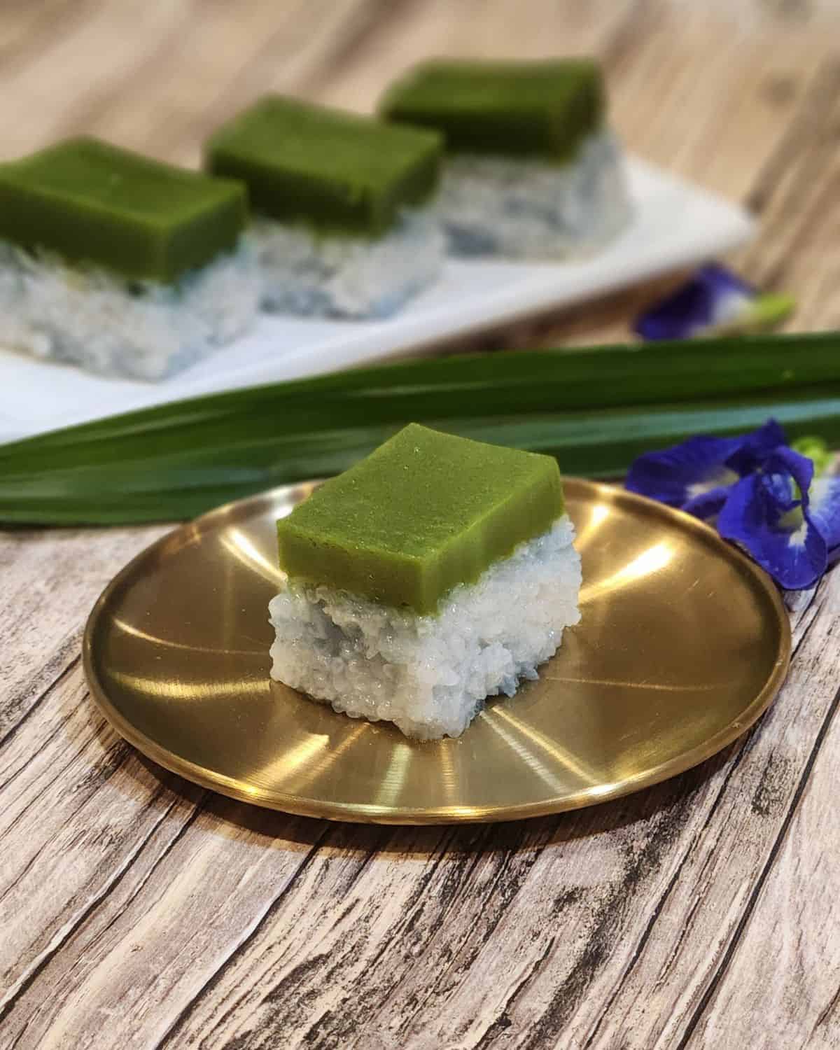 A golden plate of kuih seri muka with butterfly pea flowers and pandan leaves at the back.