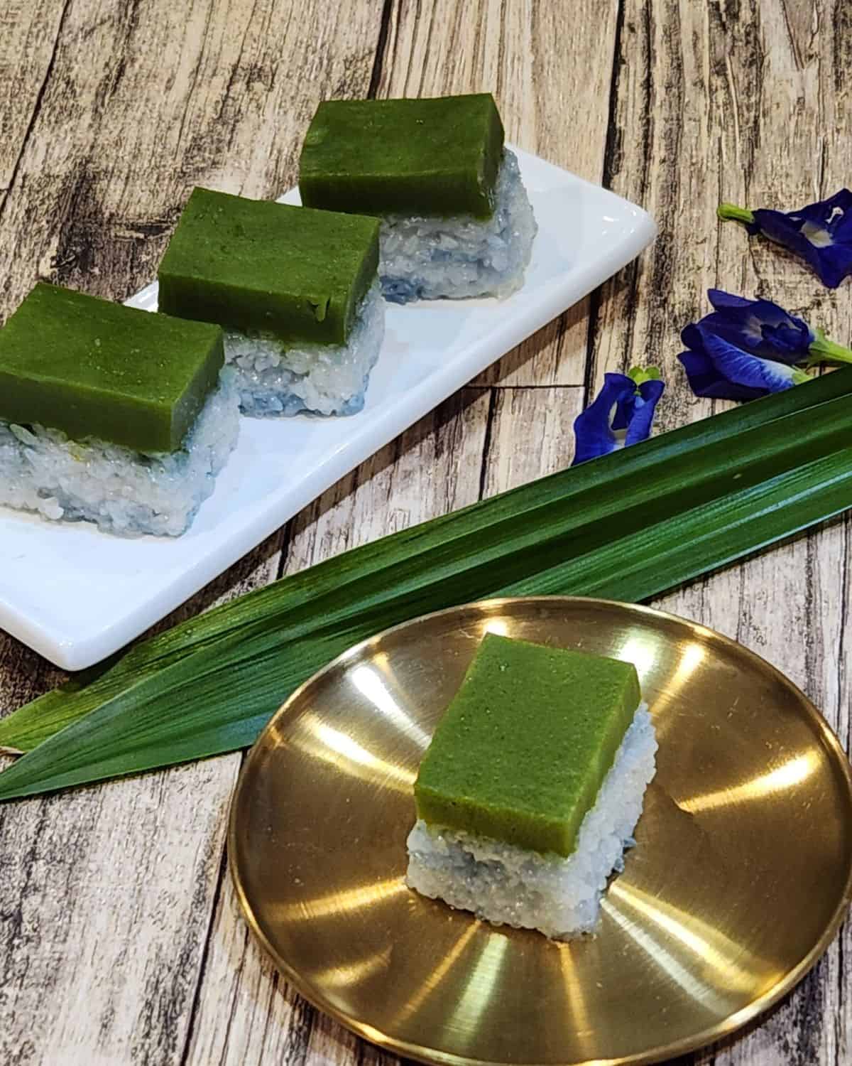 A golden plate of kuih seri muka with butterfly pea flowers and a few slices more on a white plate.