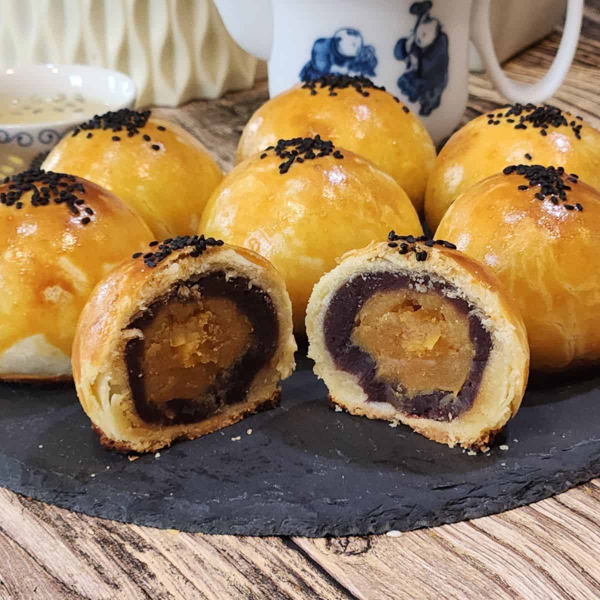 Six salted egg yolk pastry with red bean filling and one sliced open on a black tray.