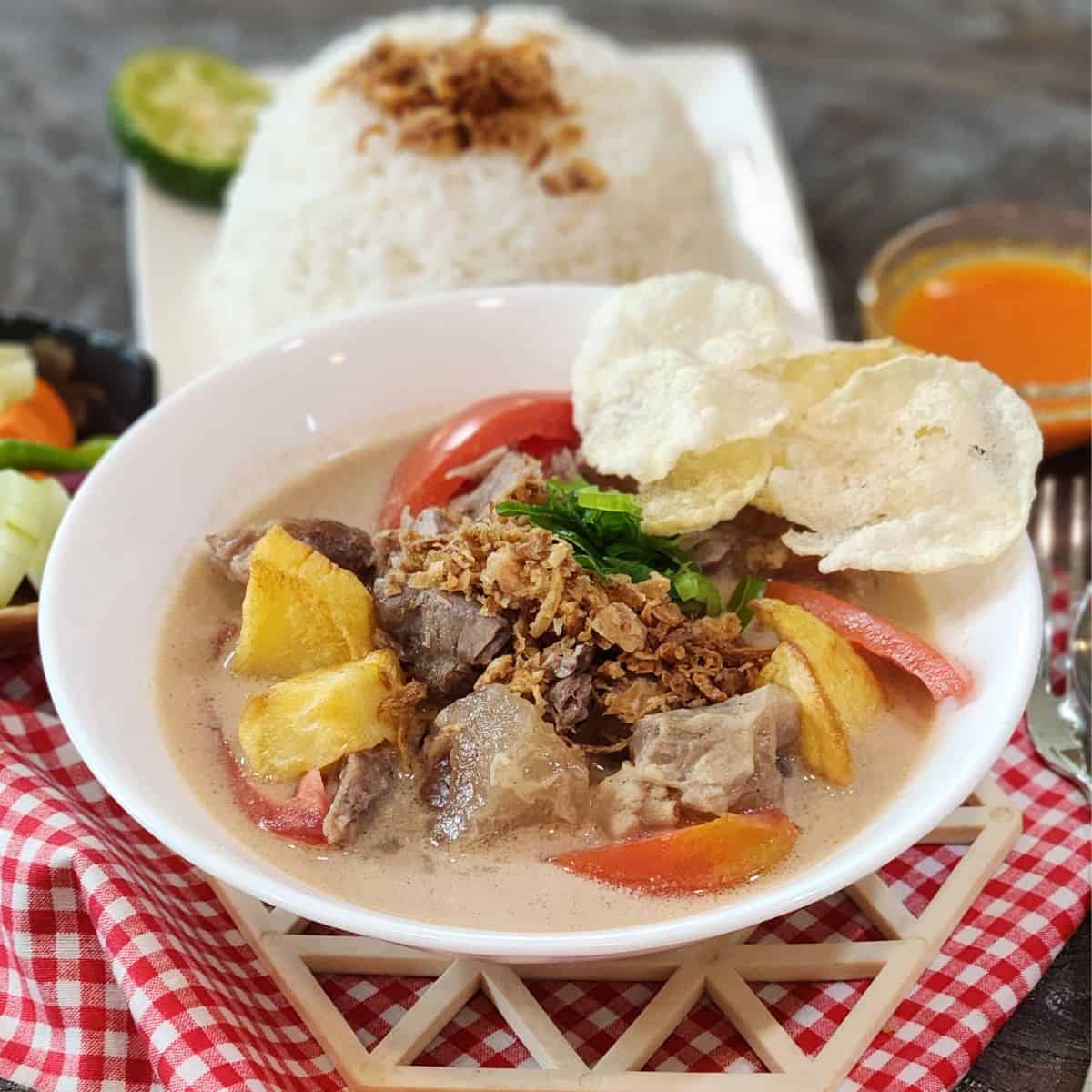 A bowl of Soto Betawi with white rice, chili sauce, and acar timun.