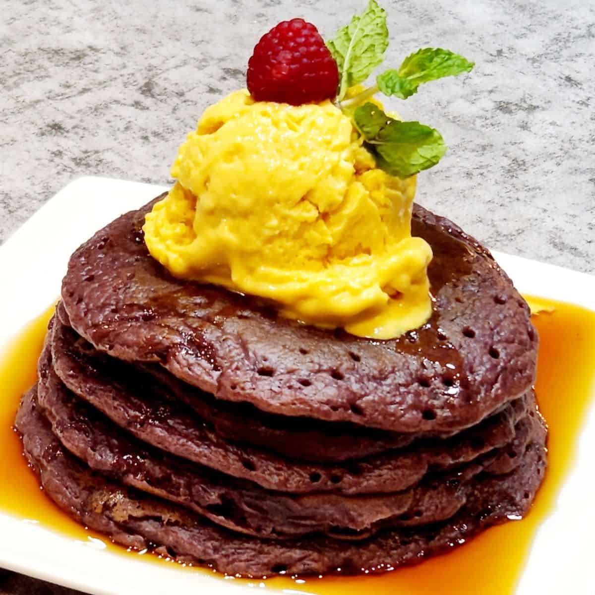 A stack of ube mochi pancakes drizzled with maple syrup and topped with mango ice cream.