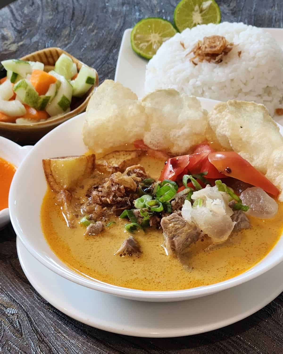 A bowl of soto tangkar, Indonesian beef coconut soup with crackers, steamed white rice, sambal, and pickles.
